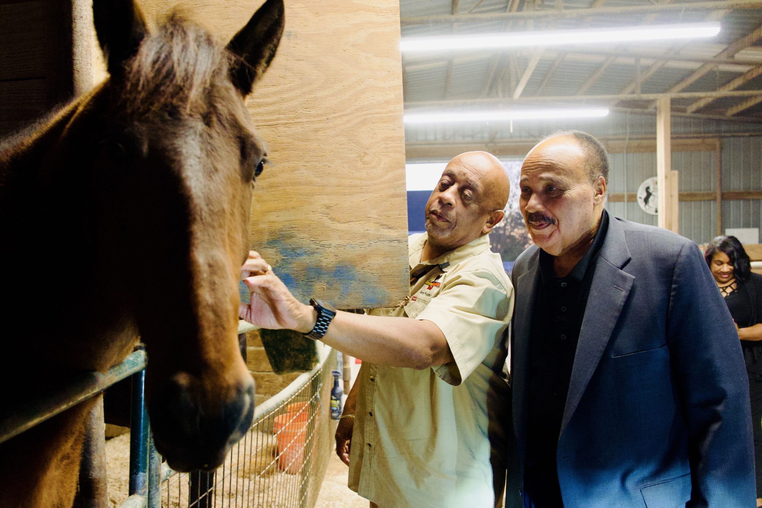 Michael Lofton visit a horse in a wooden stall, extending his hand toward the horse while and Martin Luther King, III stands beside him.