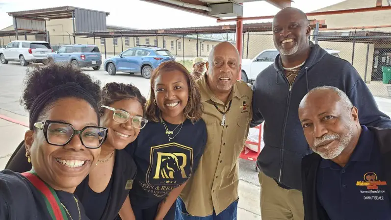 AAYHF CEO Michael Lofton with five smiling adults gather for a casual group photo beneath a metal shelter, with cars and residential-style buildings behind them.