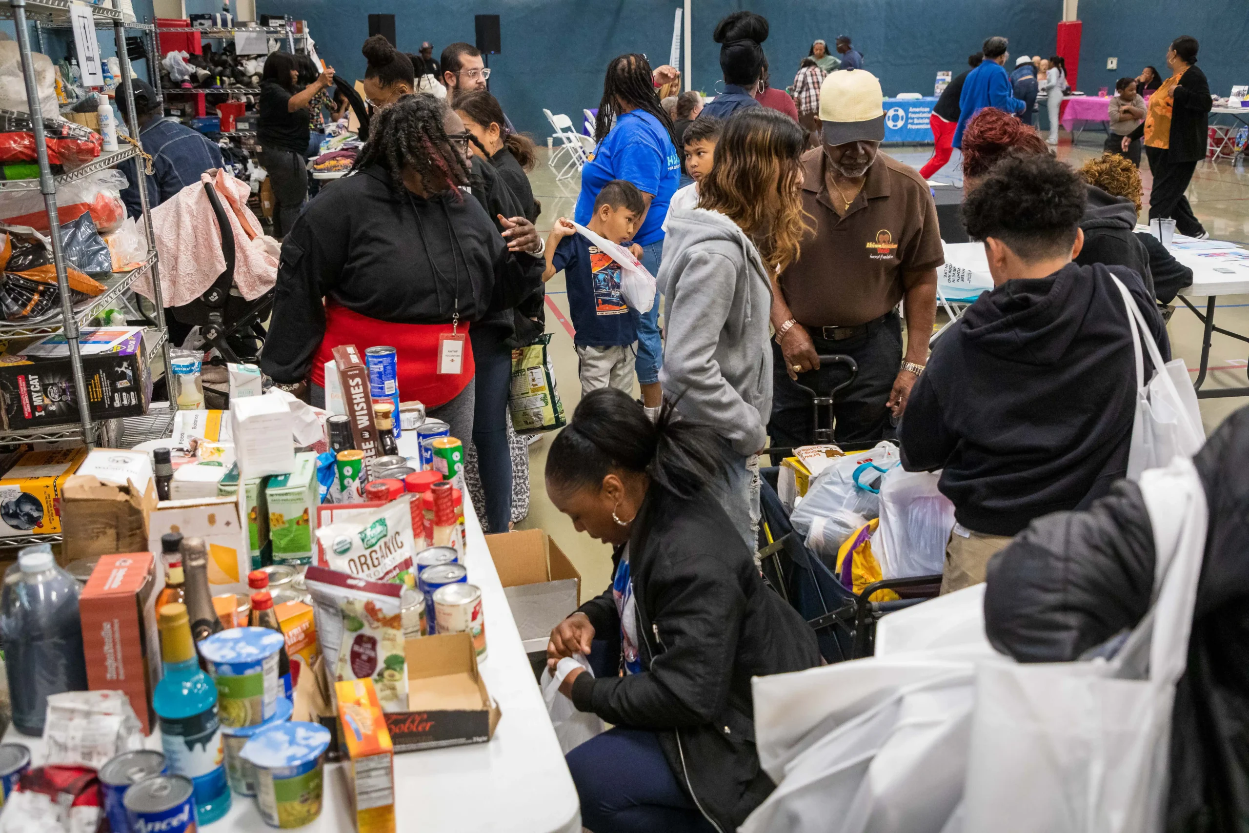A busy community giveaway takes place in a gymnasium, where adults and children browse tables filled with canned goods, pantry items, and other donated supplies.