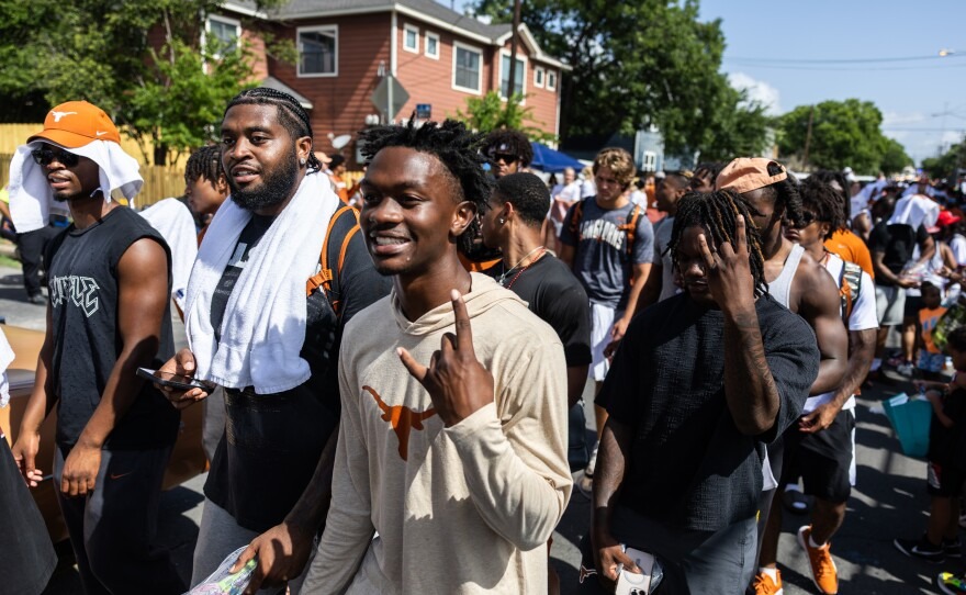A group of young people walks in the Austin Juneteenth Parade on a sunny residential street, several wearing towels over their shoulders or heads, with one smiling and gesturing toward the camera.
